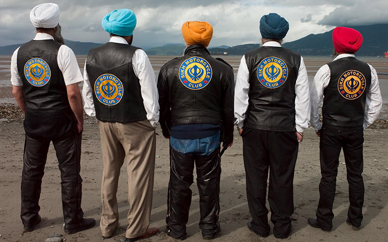 Six hommes se tiennent dos à la caméra, portant des gilets arborant l’inscription « Sikh Motorcycle Club ».