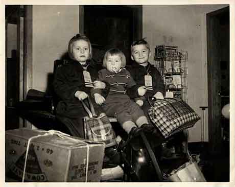 ieille image en noir et blanc sur laquelle se trouvent trois enfants assis avec des sacs de voyage. Ils se tiennent les uns les autres et semblent très heureux.