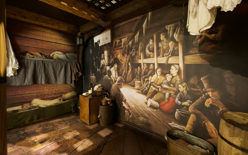 Une salle de maquettes montrant l’intérieur d’un vieux navire, avec plusieurs accessoires et une grande photo de personnes dans des couchettes.