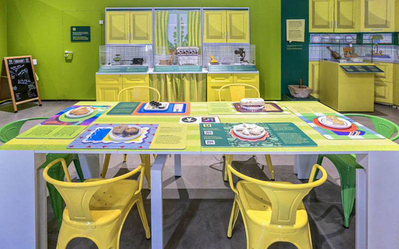 Une cuisine verte avec une table sur laquelle sont posés de faux aliments dans des assiettes, notamment du pain bannique, des sushis et des pierogis.