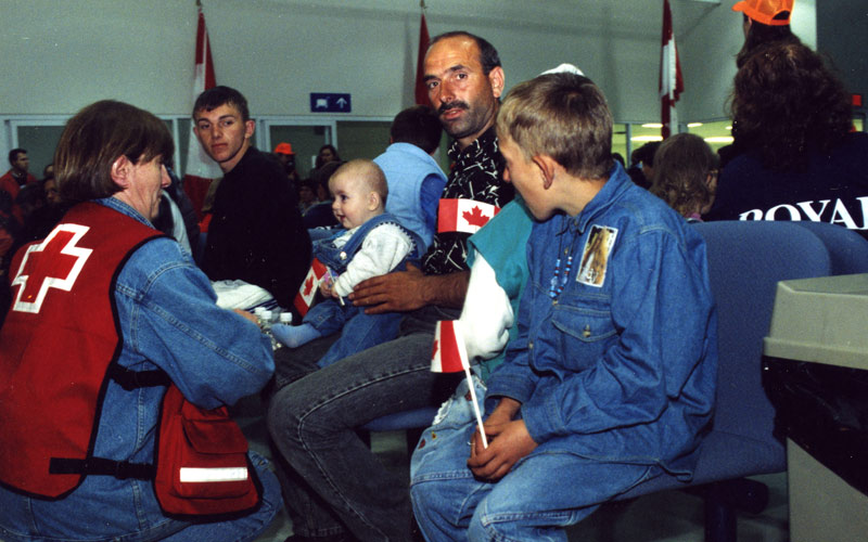Une femme blanche portant un gilet de la Croix-Rouge s’agenouille devant une famille de réfugiés kosovars composée d’un homme, de deux adolescents et d’un bébé.