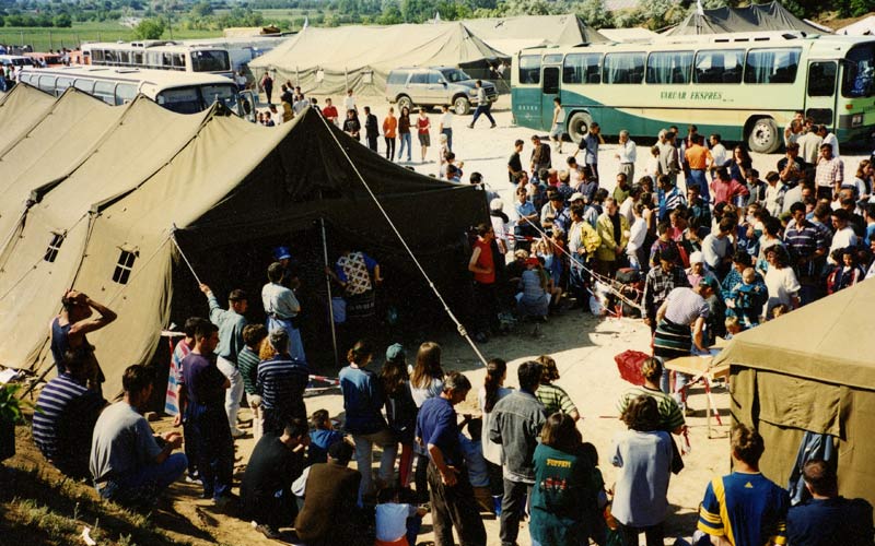 De grands groupes de réfugiés kosovars attendent à l’extérieur de tentes militaires, avec des bus en arrière-plan.