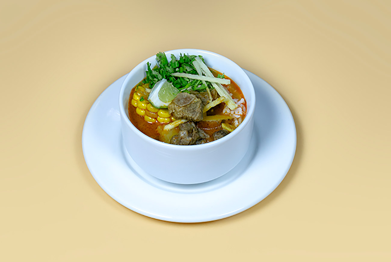 Un bol d'aloo birria avec un bouillon rougeâtre, des épis de maïs, des morceaux de bœuf, du riz, de la coriandre, des bandes de tortilla et un quartier de citron vert.
