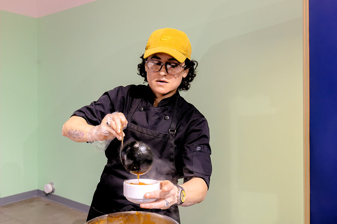 Elvira, vêtue d’un uniforme noir de chef cuisinière, verse d'aloo birria fumant dans un bol blanc.