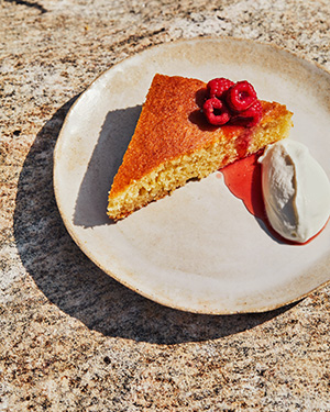 Un gâteau sur une assiette beige, garni d’une poignée de framboises sur le dessus et de crème fouettée au yogourt sur le côté.