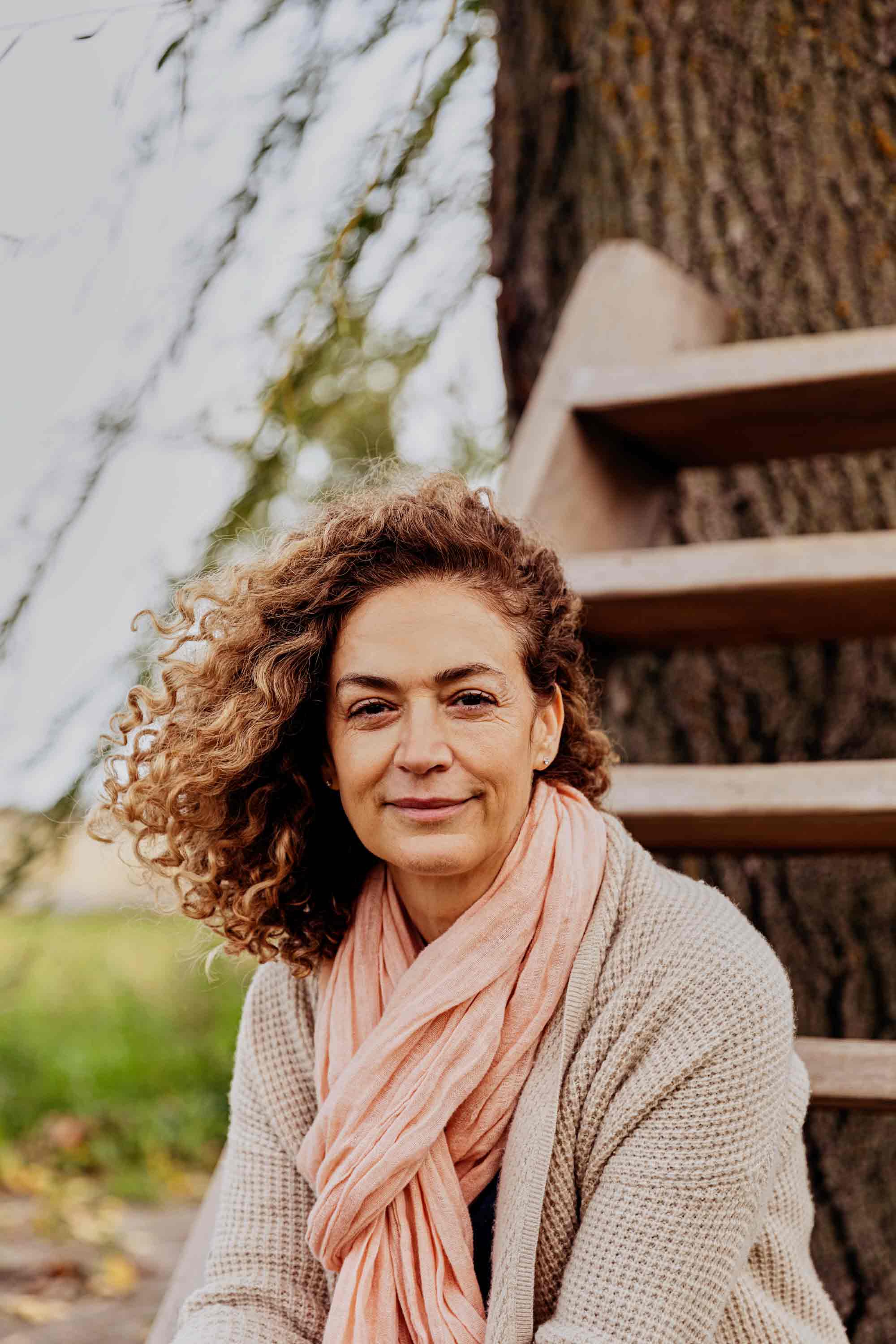 Portrait d’une femme turque souriante, les cheveux bouclés sur le côté, portant un foulard rose et un cardigan beige.