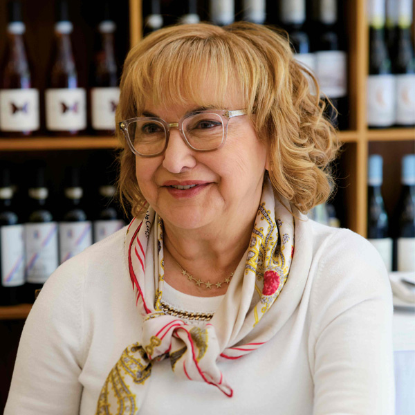 Portrait d’une femme italienne souriante portant un foulard de soie et une chemise blanche, avec des bouteilles de vin en arrière-plan.