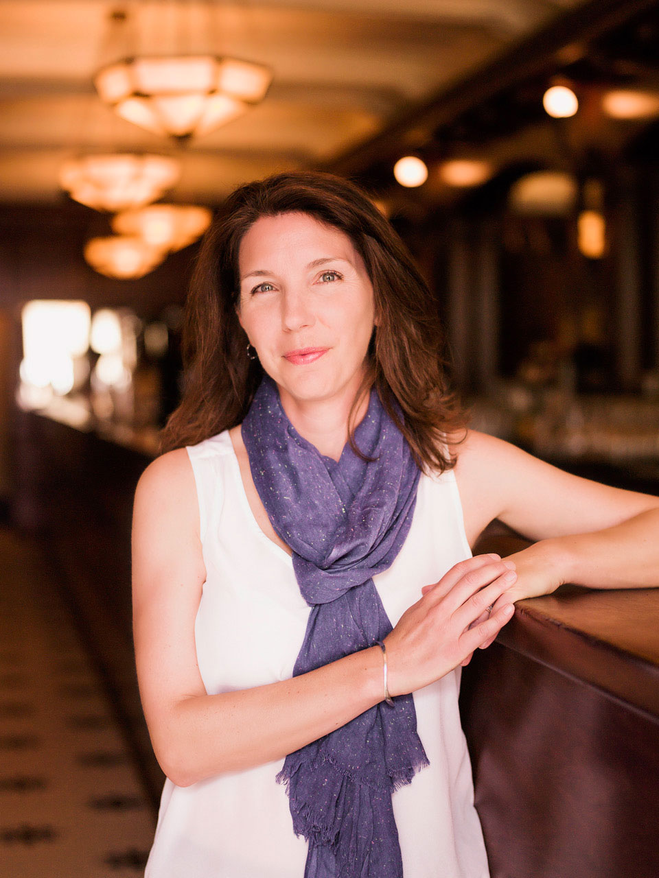 Portrait d’une femme blanche souriante, vêtue d’une camisole blanche et d’un foulard violet, accoudée à un bar.