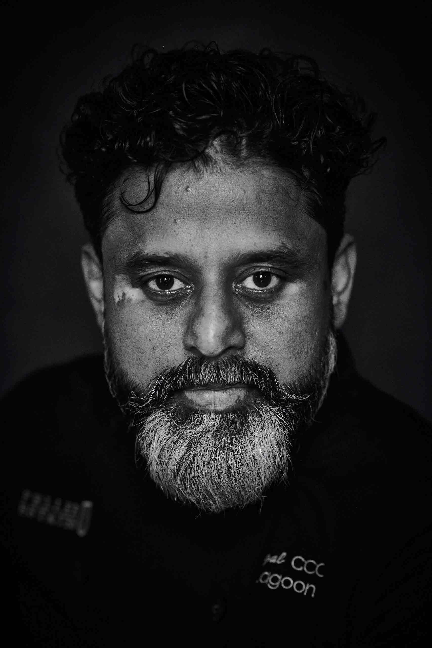 Portrait en noir et blanc d’un homme indien aux cheveux courts et bouclés et à la barbe grisonnante qui regarde l’appareil photo.