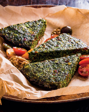 Trois morceaux de kookoo sabzi servis dans une assiette avec du papier parchemin, sur une table en bois.