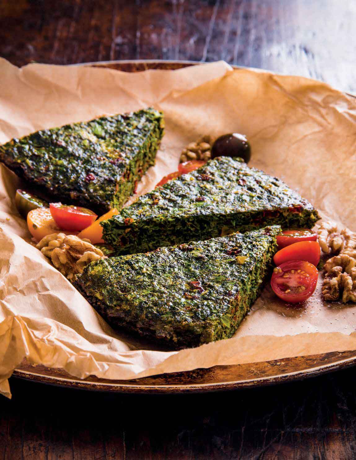 Trois morceaux de kookoo sabzi servis dans une assiette avec du papier parchemin, sur une table en bois.