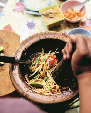 Salade de papaye dans un mortier brun, mélangée avec un pilon et une cuillère de service, avec les ingrédients à l’arrière-plan.