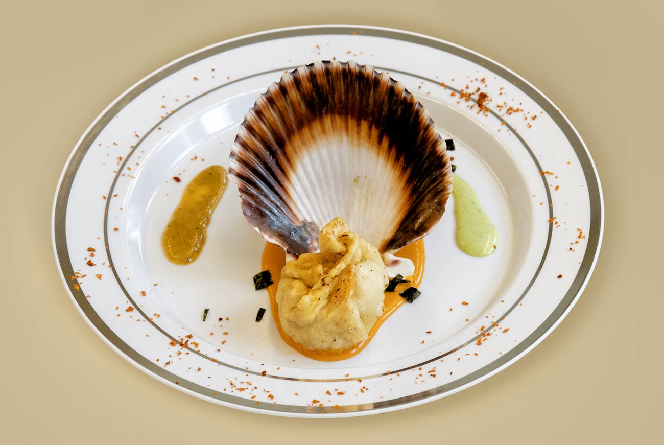 Un momo aux fruits de mer placé devant une coquille Saint-Jacques dans une assiette blanche, sur un fond de sauce orange, avec une sauce ail et coriandre à gauche et un chutney de timut à droite.