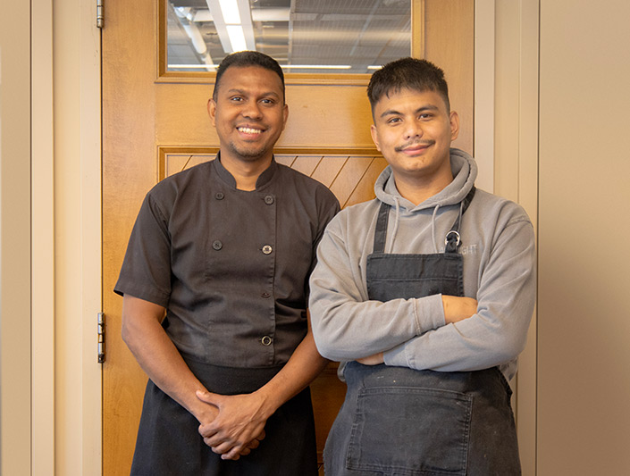 Deux chefs, Darwin et Rohit, sont debout, souriants, devant un mur et une porte en bois.