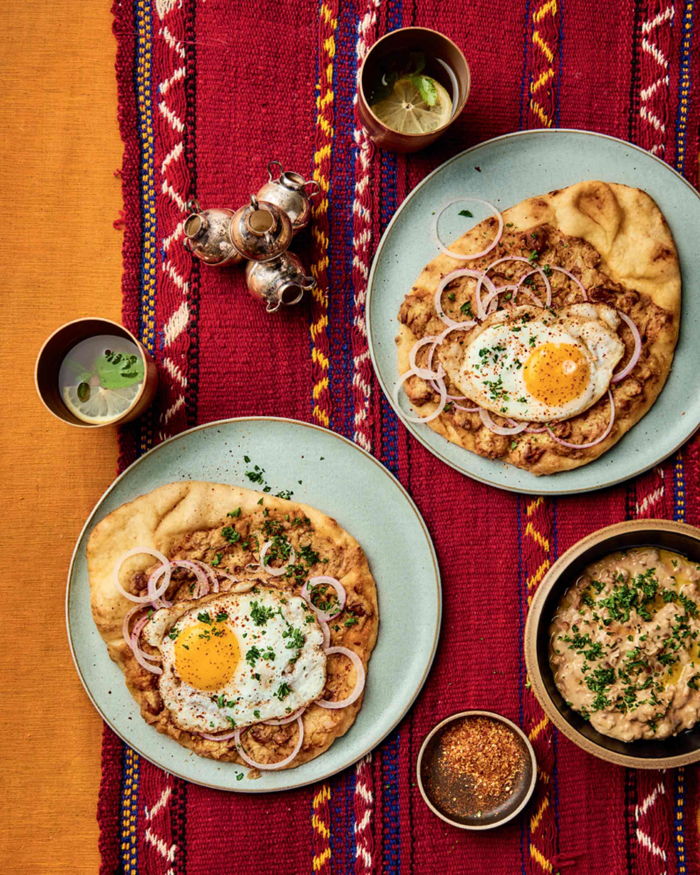 Deux assiettes de pita, d’œufs et d’oignons, des mudammas complets dans un bol et deux boissons dans des tasses, le tout posé sur un tissu rouge décoré.