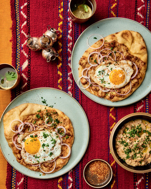 Deux assiettes de pita, d’œufs et d’oignons, des mudammas complets dans un bol et deux boissons dans des tasses, le tout posé sur un tissu rouge décoré.