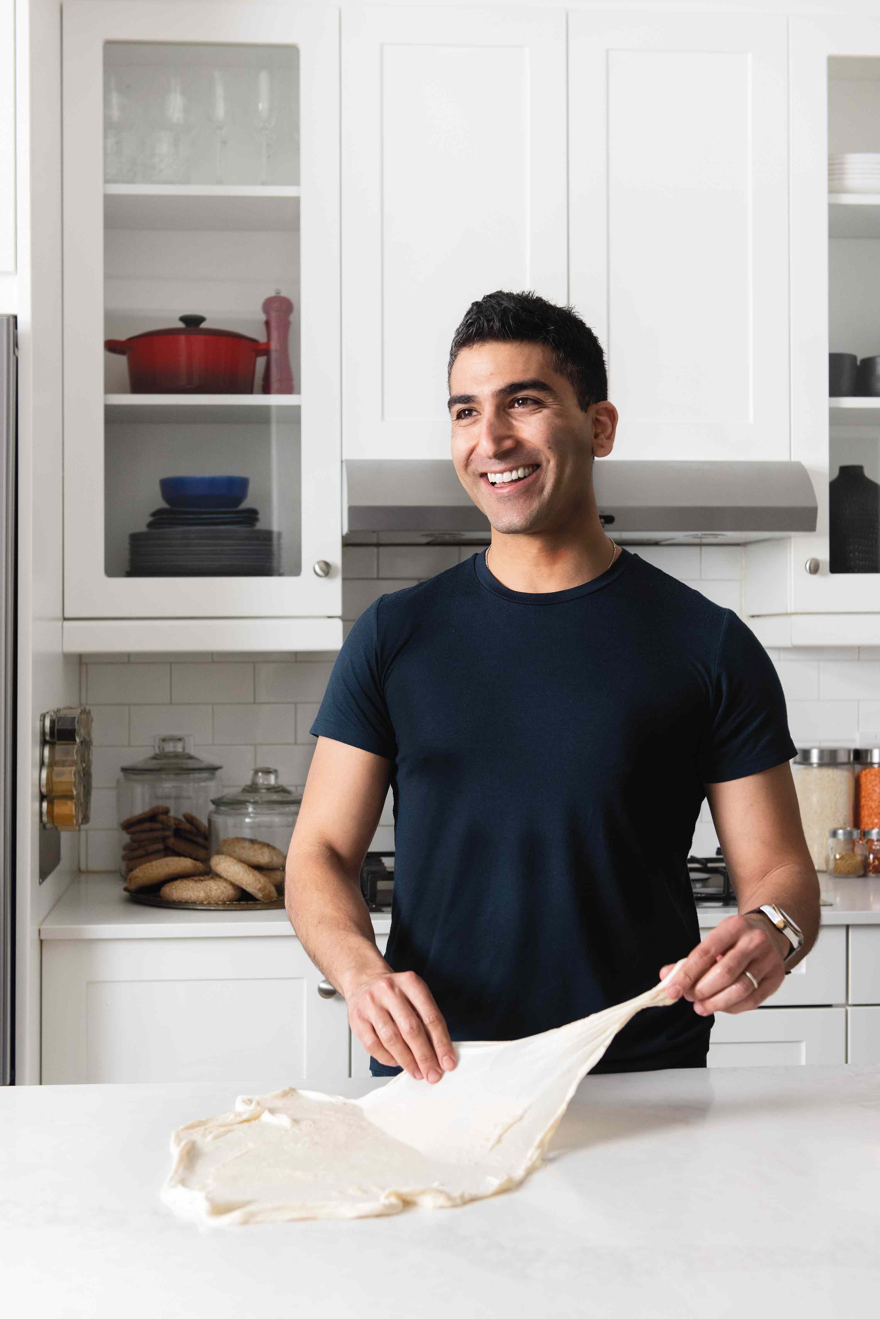 Un jeune homme souriant d’origine égyptienne plie de la pâte dans une cuisine blanche et lumineuse.