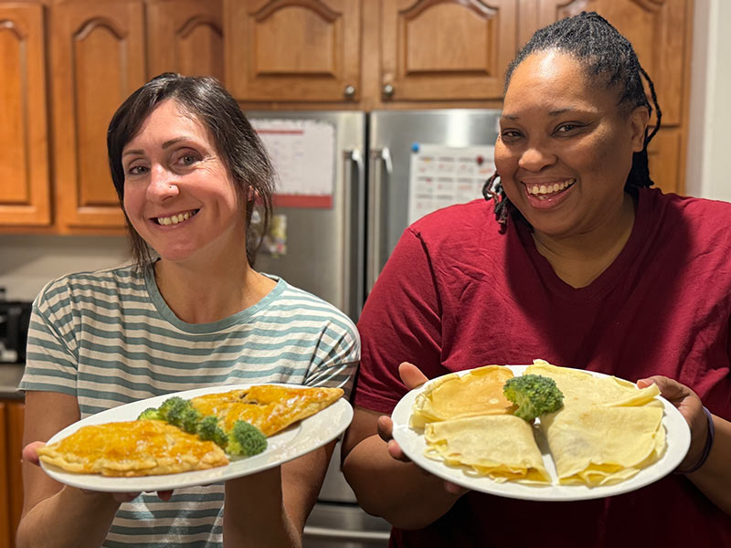 Les chefs Celia et Karyna sourient chaleureusement dans une cuisine en présentant des Ukajas fraîchement préparés sur de grandes assiettes blanches.