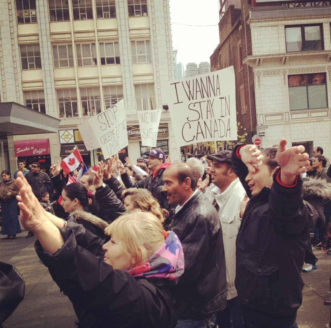 Une foule de personnes manifestent dans une rue de la ville en brandissant des pancartes, sur une pancarte, on peut lire Je veux rester au Canada.
