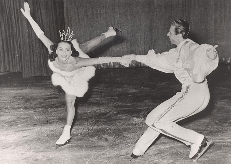 Photo en noir et blanc d’un homme faisant du patinage artistique avec une femme qui porte une petite couronne, la jambe en l’air.