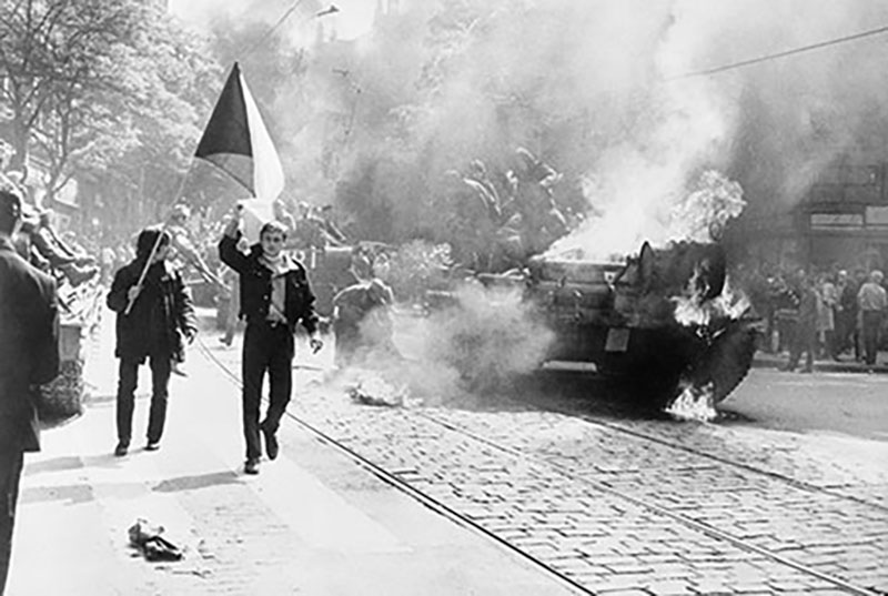 Photo en noir et blanc de personnes marchant dans une rue, une personne agite un drapeau, un tank en arrière-plan semble être en feu.