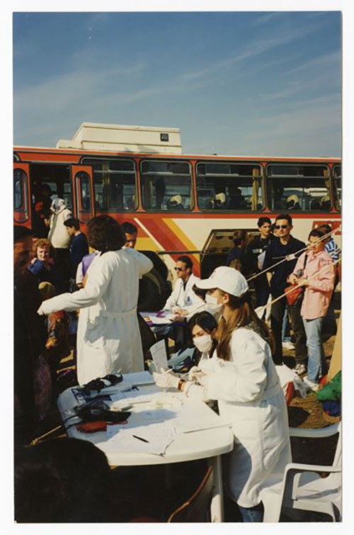 Des gens descendent d’un autobus alors que deux femmes assises à une table portent des masques médicaux blancs.