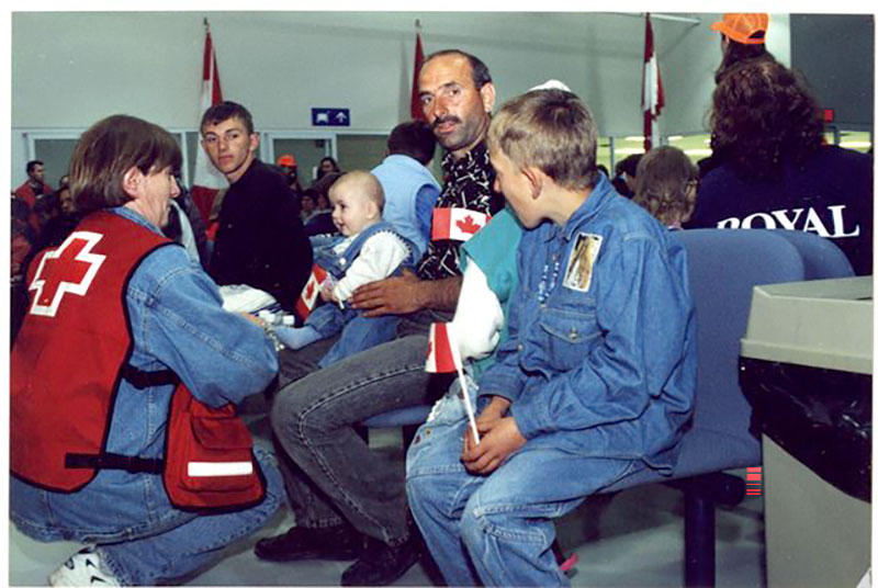 Un homme assis, tenant un bébé sur ses genoux. Le bébé tient un petit drapeau canadien et sourit à une femme portant un gilet de la Croix-Rouge.