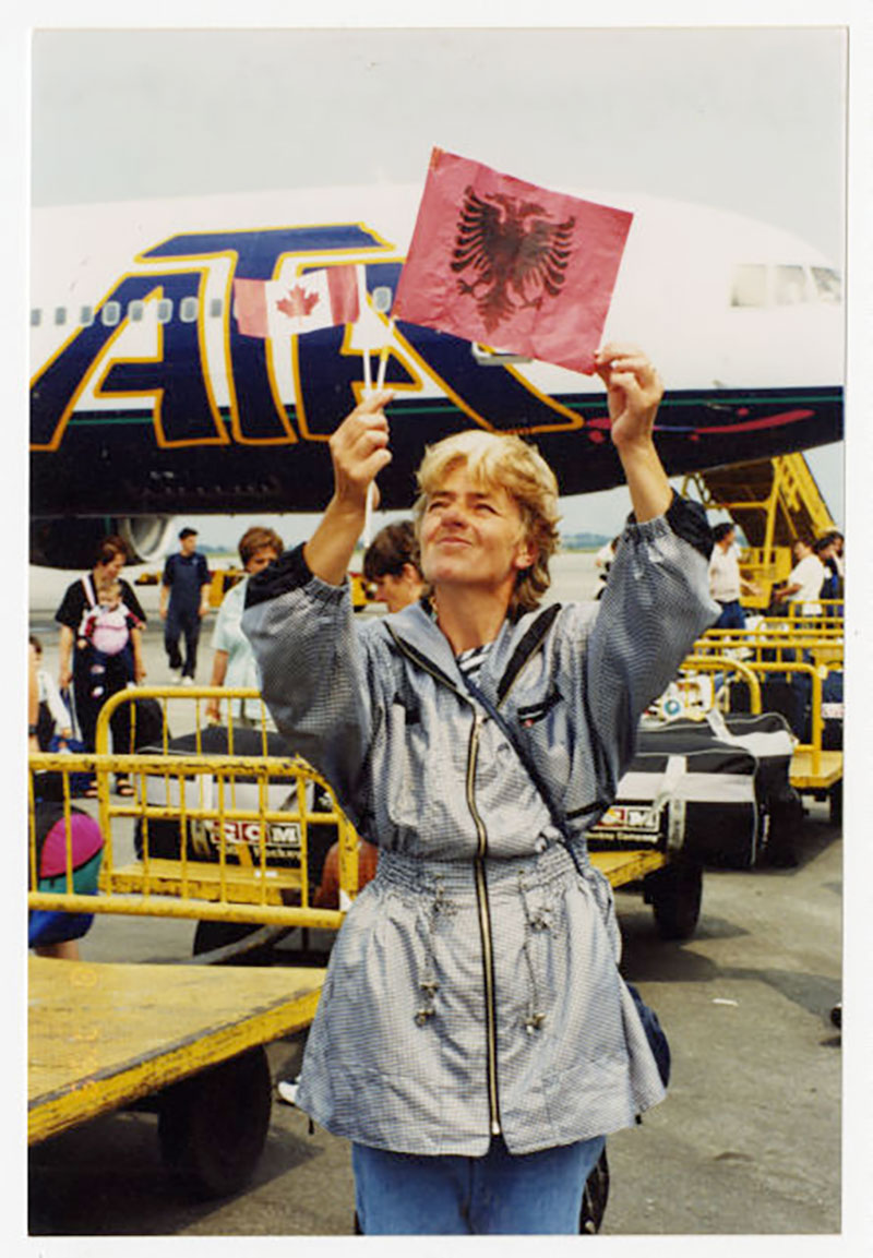Une femme tient un carré de papier rouge sur lequel est affiché un emblème noir. Un avion se trouve derrière elle.