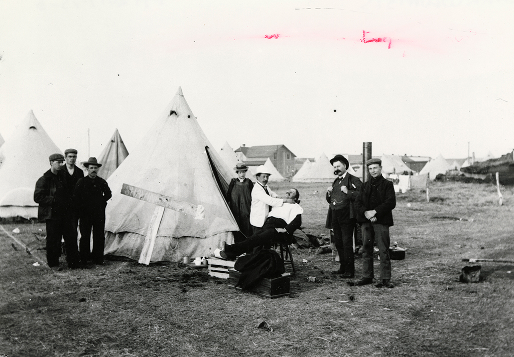 Plan rapproché de l’une des centaines de tentes dans un camp de travail; des hommes et des femmes se tiennent à côté de la tente pendant qu’un barbier prépare un homme à se raser.