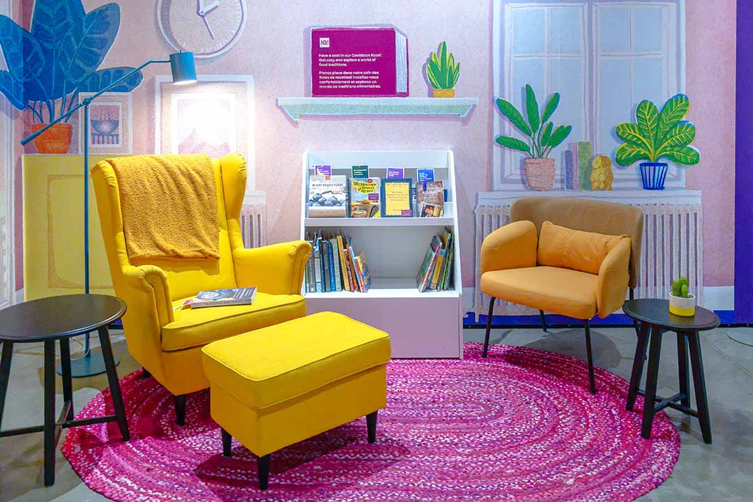 Dans une pièce aux murs roses, une chaise et un pouf jaunes sont posés sur un tapis rose à côté d’une étagère contenant deux douzaines de livres de cuisine.