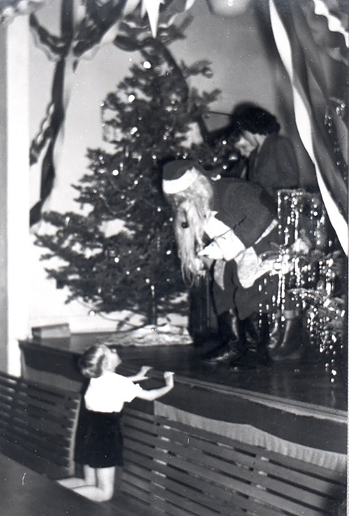 Un homme habillé en Père Noël sur une scène à côté d’un arbre de Noël. Il se penche pour parler à un garçon sur le plancher.