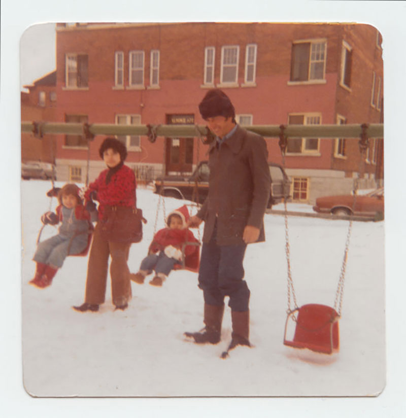 Photo vintage d'un homme et d'une femme poussant chacun un enfant sur une balançoire. La neige recouvre le sol et ils sont devant un grand bâtiment en briques.