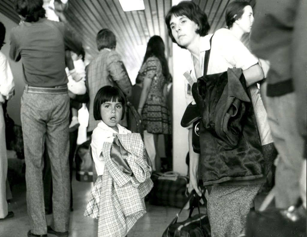 Image en noir et blanc d’une enfant aux cheveux foncés qui regarde la caméra avec son manteau sur le bras. À côté d’elle, une femme aux cheveux foncés a son manteau sur le bras et tient un sac de voyage. Elles sont entourées d’autres personnes qui tournent le dos à la caméra.