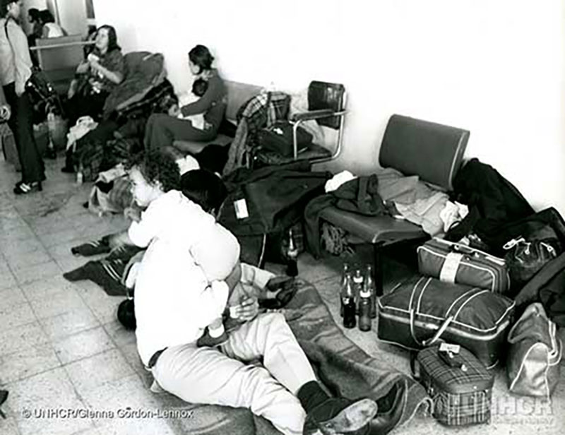 Image en noir et blanc d’une pièce avec une plancher de tuiles et une rangée de chaises le long du mur. Des vêtements et bagages sont éparpillés et un homme est assis sur une couverture étendue sur le plancher, avec un bébé sur l’épaule.