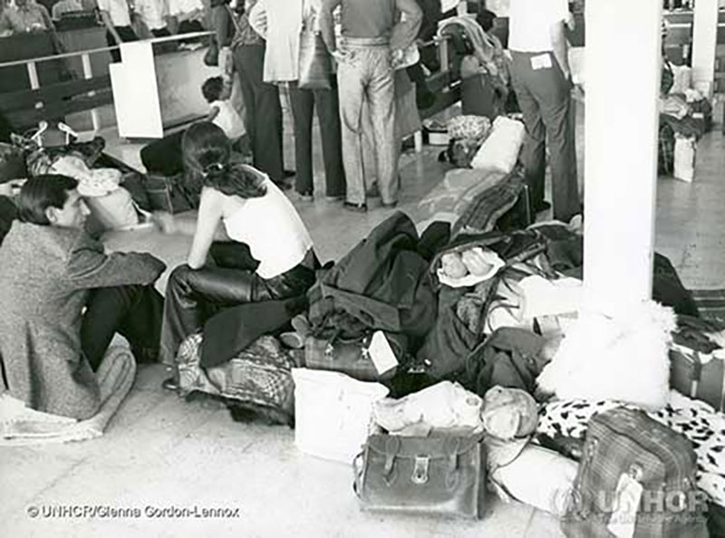 Photo en noir et blanc d’une homme et d’une femme assis sur un plancher de tuiles à côté d’une pile de bagages et de vêtements. En arrière-plan, d’autres personnes tournent le dos à la caméra et il y a d’autres bagages éparpillés sur le plancher.