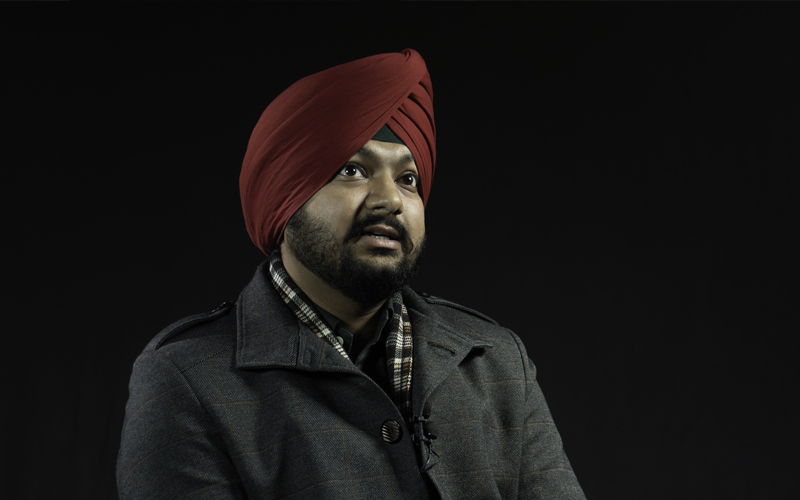Homme sud-asiatique avec un turban, une barbe, un veston gris avec des lignes rouges et un foulard sur fond noir.