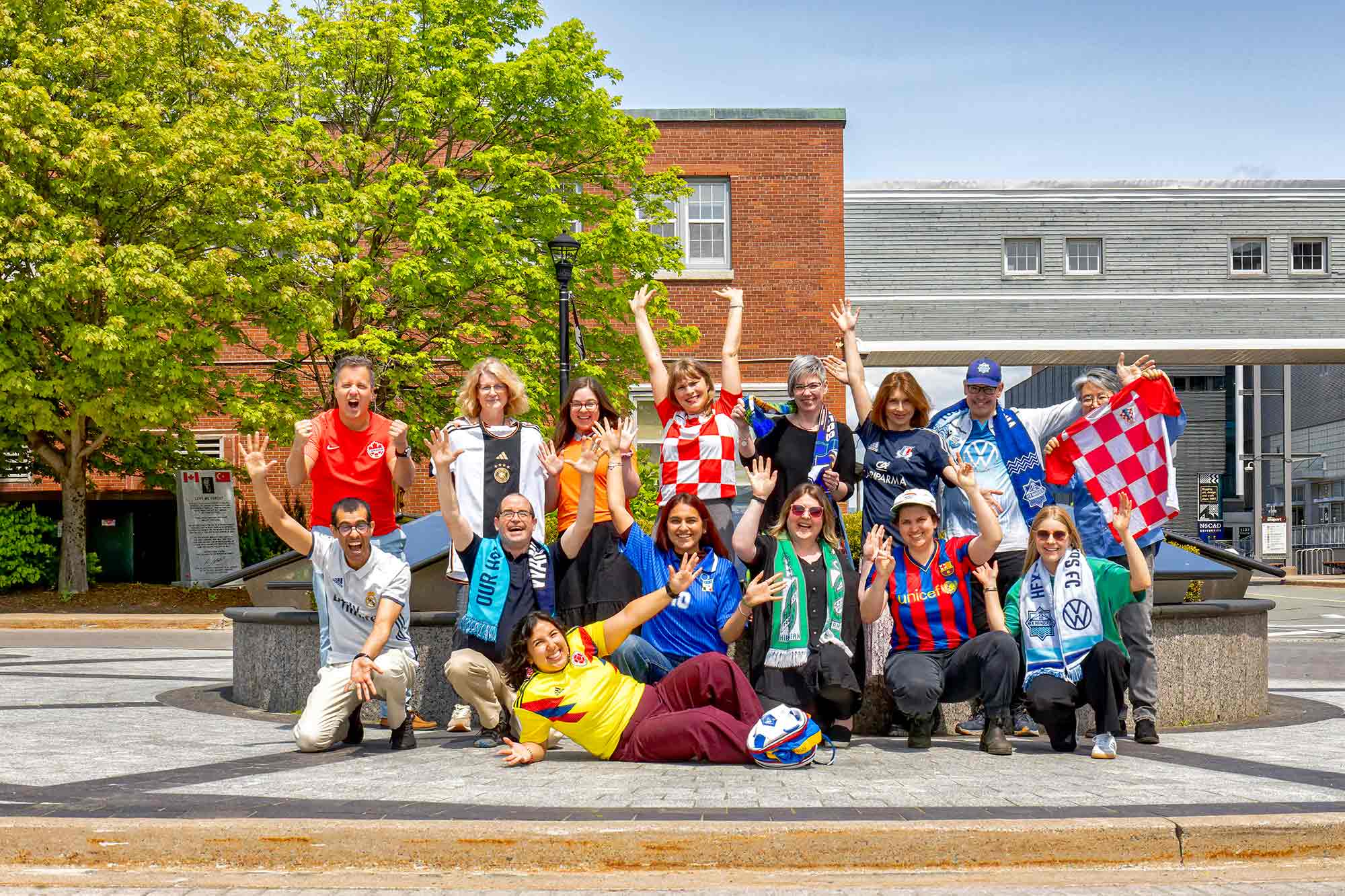 Quinze personnes applaudissant, portant les couleurs de différentes équipes de soccer, ils forment deux rangées, et une femme est allongée sur le sol devant eux. 