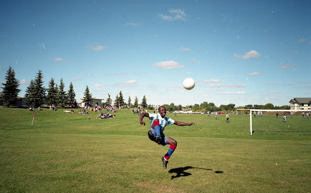 Un homme noir dont les pieds ne touchent plus le sol sourit après avoir botté un ballon. Il est en plein soleil, sur un grand terrain, devant terrain de soccer en activité.