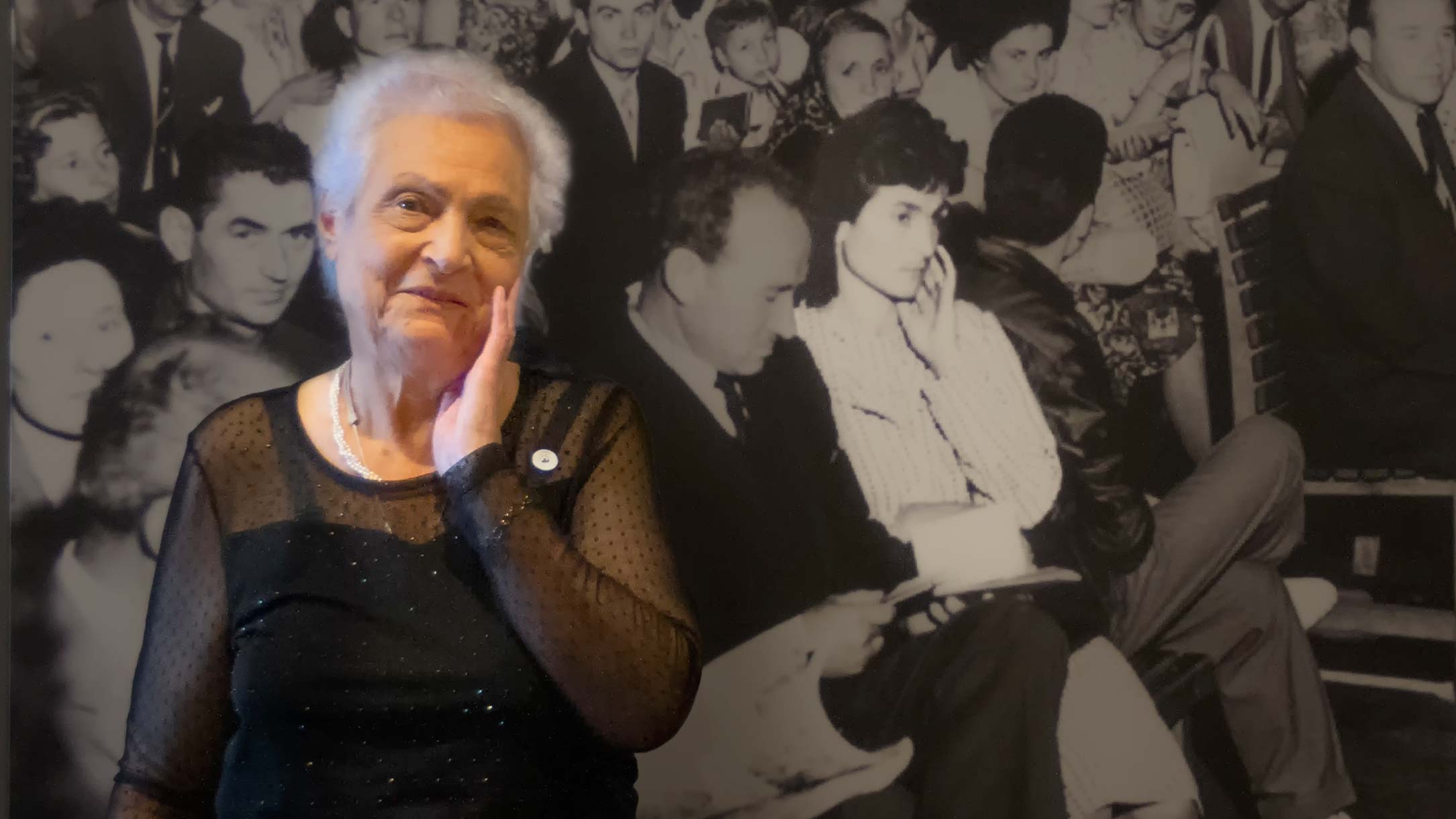 Une femme âgée se touche le visage devant une grande photographie en noir et blanc. Sa pose reflète le geste de la jeune femme sur la photo ― sa propre image, lorsqu’elle était jeune.