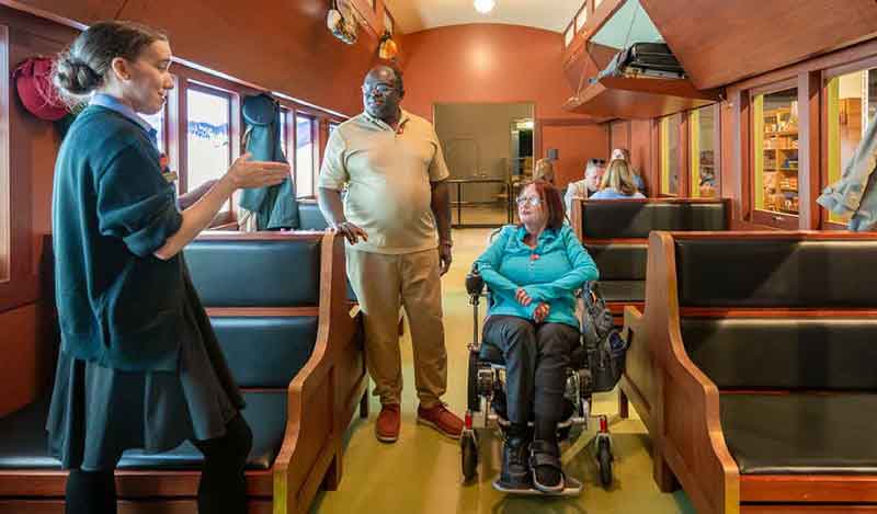 Un guide du Musée s’entretient avec deux visiteurs, dont l’un est en fauteuil roulant, à l’intérieur d’une exposition présentant un wagon historique équipé de banquettes en bois.