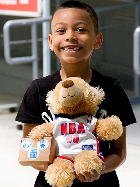 Un jeune garçon souriant tient un ourson habillé comme un joueur de basketball.