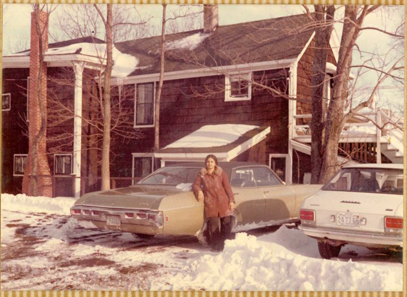 Une jeune femme s’appuie sur une voiture dans son allée, il y a beaucoup de neige sur le sol.