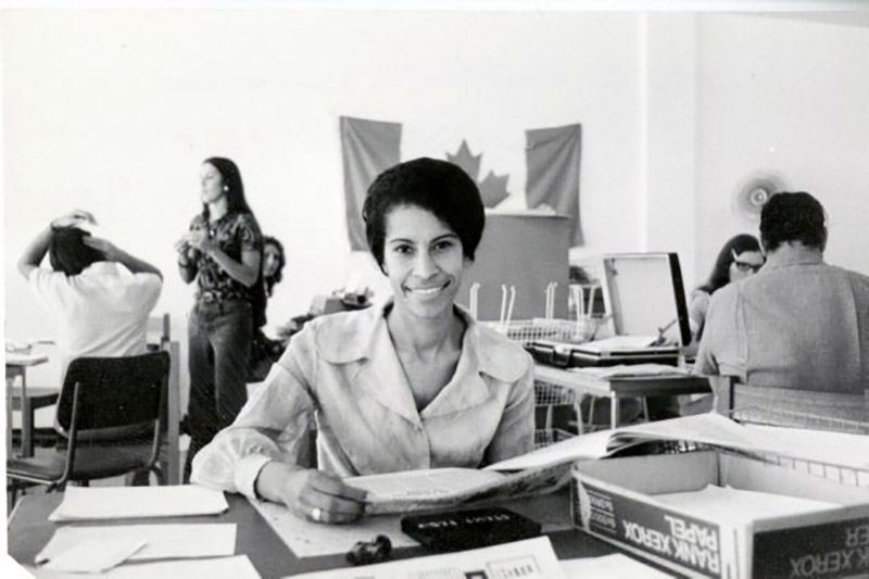 Une jeune femme assise à un bureau sourit et regarde des documents. Le drapeau canadien est accroché au mur derrière elle.