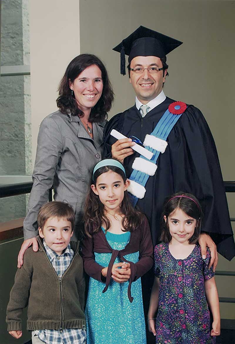Un jeune homme portant la toge et le mortier tient fièrement son diplôme en compagne de sa femme et de ses trois enfants.