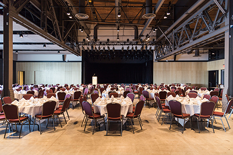 Une grande salle dans laquelle se trouvent plusieurs tables à manger rondes recouvertes de nappes blanches. Il y a six chaises rouges autour de chaque table.