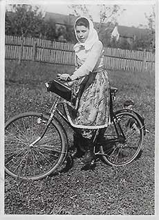 Une photographie en noir et blanc d'une femme portant une jupe à fleurs et un fichu. Elle est debout, près d'un vélo