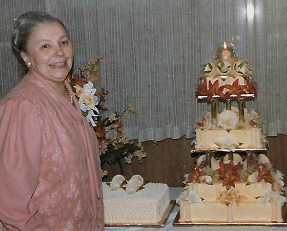 Une femme portant une robe rose sourit près d'un gâteau à trois étages qui est décoré de façon élaborée