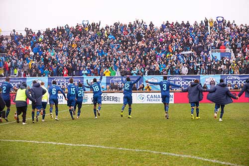 Une grande foule de fans enthousiastes occupent les gradins. Sur le terrain, une ligne de joueurs de soccer en uniformes bleus lèvent la main vers la foule.