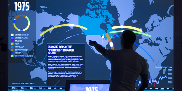Une personne, en silhouette, montre une grande carte du monde projetée avec des lignes qui relient différentes régions au Canada.