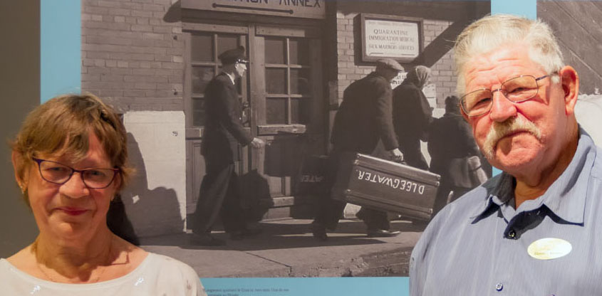 Un homme et une femme âgés se tiennent de part et d’autre d’une photo murale imprimée en noir et blanc d’un homme portant une valise sur laquelle est peint le nom Leegwater.